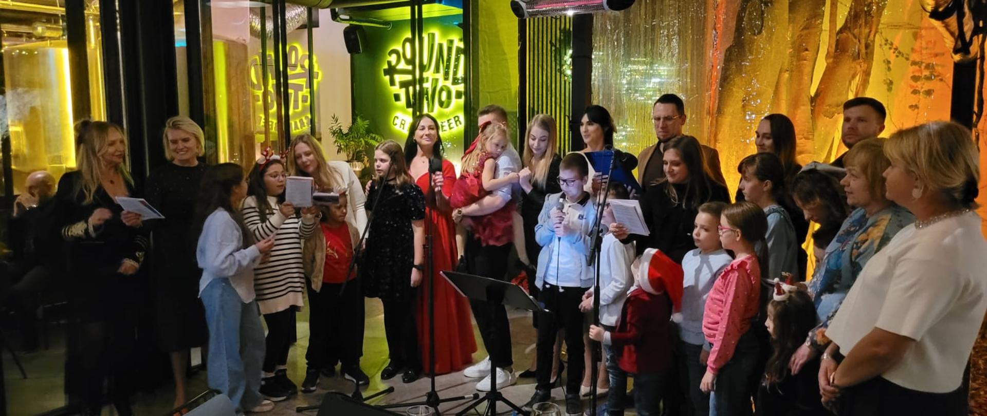 Grupa ludzi w śpiewająca do mikrofonów w lokalu. Wśród nich dorośli, młodzież i dzieci.