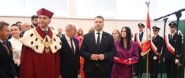 Na sali stoi mężczyzna w czerwonym ceremonialnym stroju z gronostajowym kołnierzem, za nim minister Czarnek i kobieta w fioletowej sukience, w tle wiele osób.