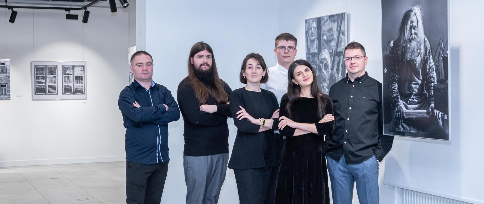 Zdjęcie przedstawia autorki i autorów wystawy fotograficznej Dni Autorskie. Stoją obok siebie w Sali w wystawienniczej. W tle wiszą czarno-białe zdjęcie. 