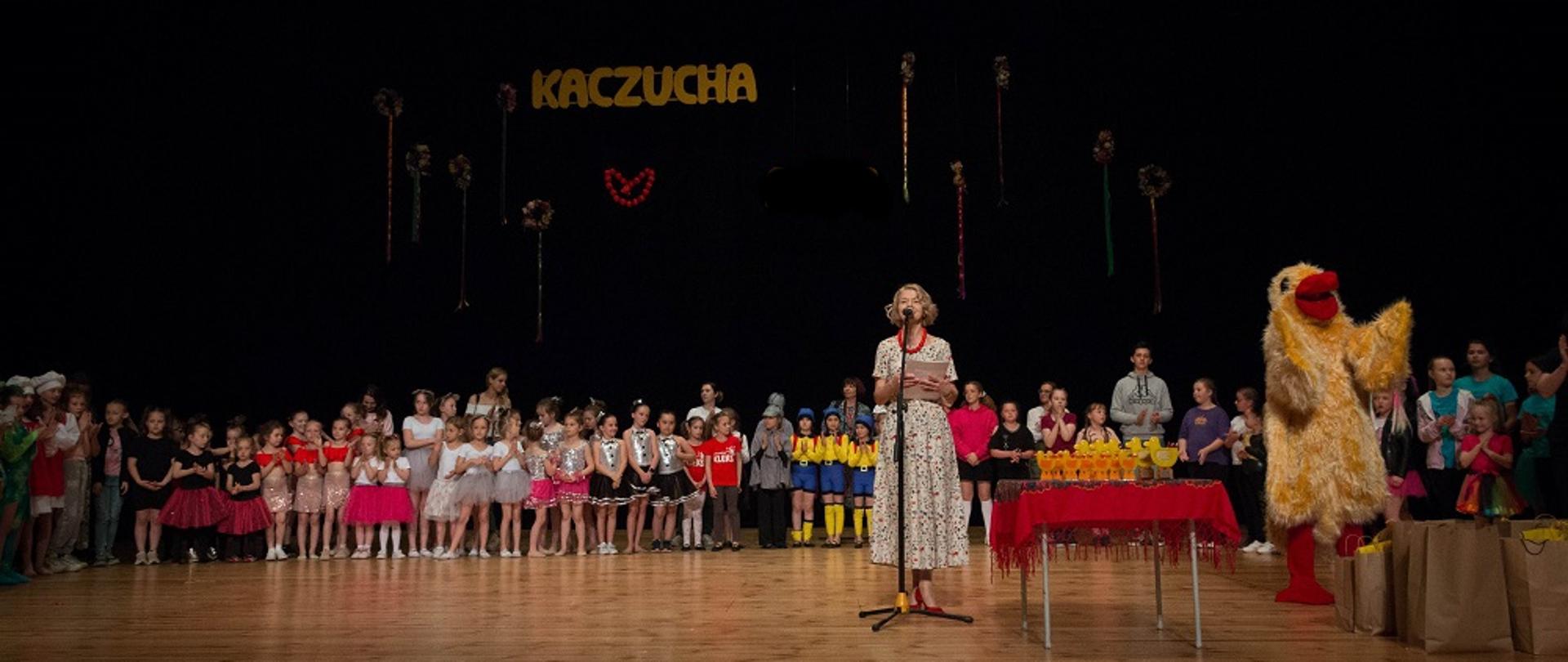 Na scenie stoi grupa dzieci podczas Przeglądu Kaczucha. Prawej stronie stoi osoba przebrana w żółty strój kaczki. Na środku stoi kobieta przemawiająca przez mikrofon.