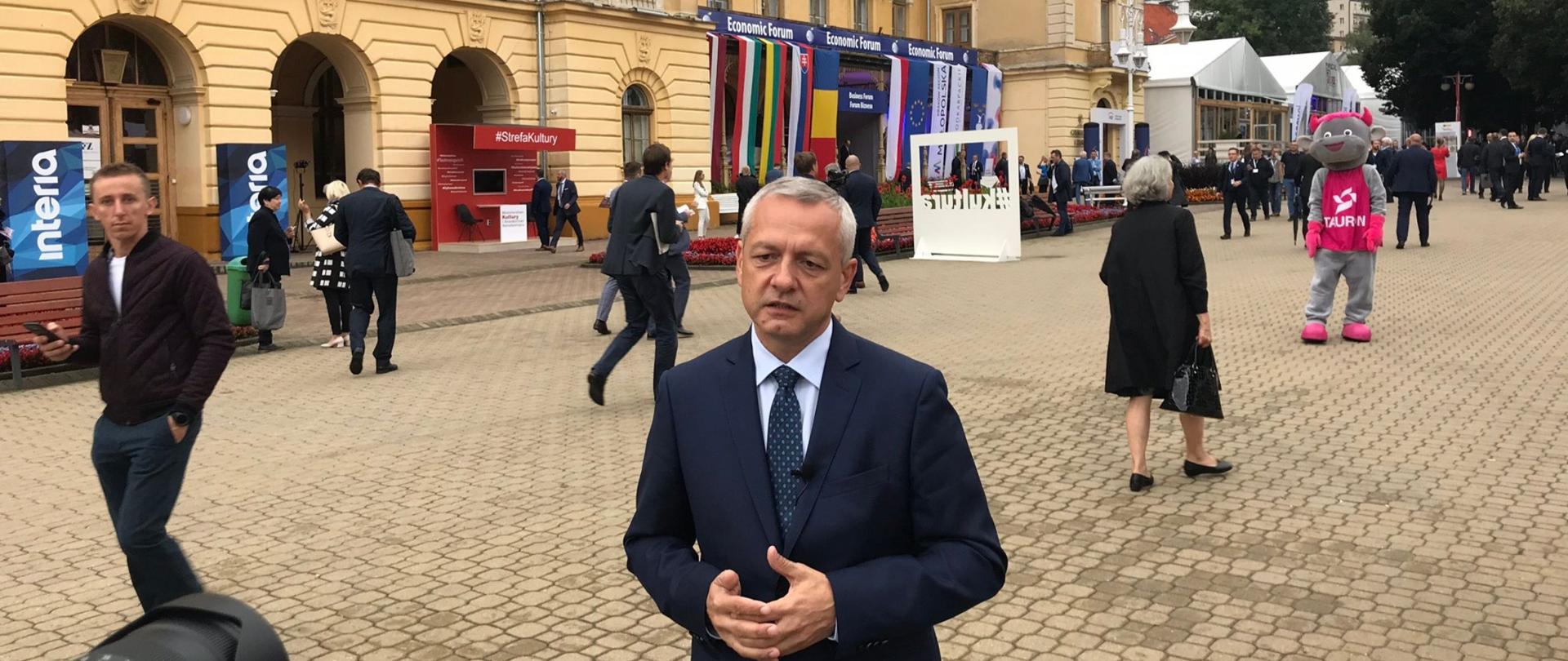 Minister Marek Zagórski na deptaku w Krynicy, w tle Stary Dom Zdrojowy i wielu przechodniów