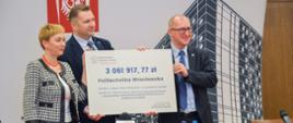 Minister Czarnek stoi za stołem i trzyma wielki symboliczny czek z napisem 3 061 917,77 zł, obok niego mężczyzna w garniturze i kobieta w ubraniu w biało-czarna kratkę.