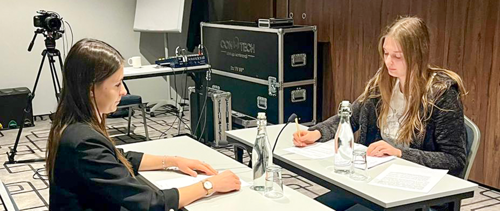 Dwie kobiety siedzą przy stolikach i patrzą się na siebie. Na stoliku leżą kartki oraz butelki z wodą.