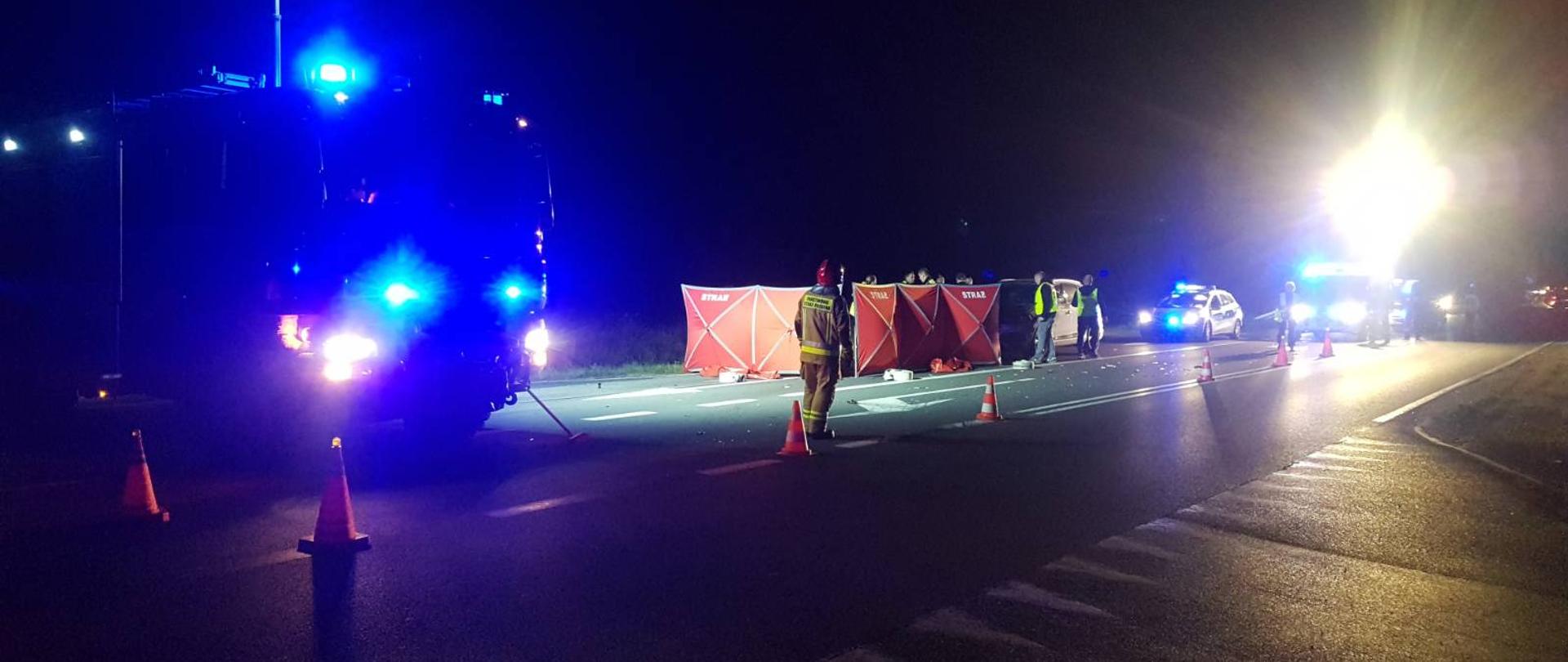 Wypadek ze skutkiem śmiertelnym. Droga wojewódzka nr 434 w okolicach Roszkówka. Na jezdni samochód, który potrącił rowerzystę, parawan ochronny oraz wozy straży pożarnej i radiowóz policyjny. Pojazdy służb mają włączone sygnały uprzywilejowania. Na jezdni stoi strażak, a za parawanem policjanci. Przy pomocy masztów oświetleniowych, strażacy oświetlają teren.