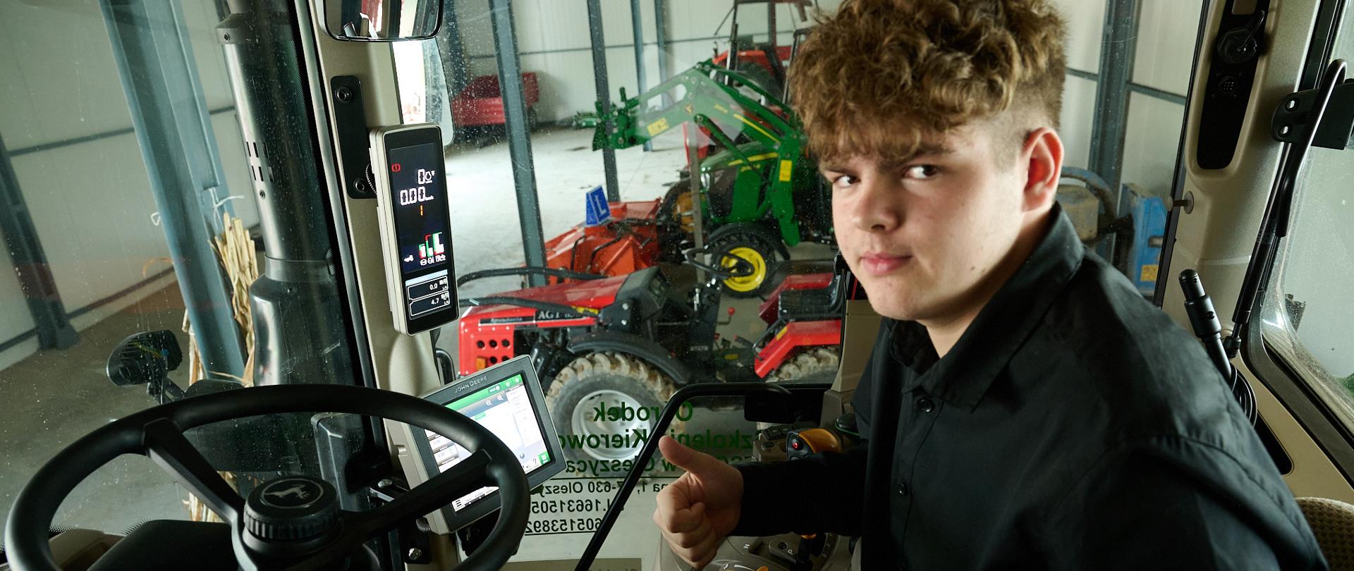 Uczeń siedzi w ciągniku rolniczym trzymając kciuk prawej ręki skierowany ku górze. W tle, z prawej strony stoją maszyny rolnicze, z lewej - przyczepa. Ciągnik i maszyny znajdują się w pomieszczeniu