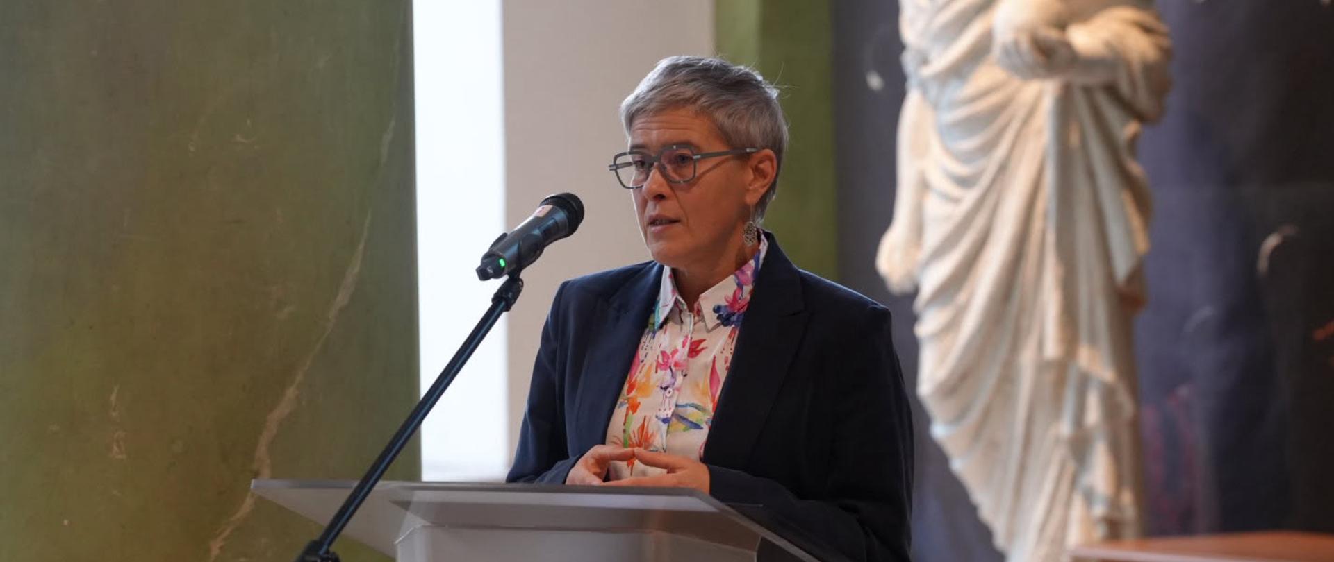 Zdjęcie z boku pod kątem, wiceminister Mrówczyńska stoi za mównicą i mówi do mikrofonu na stojaku, za nią biała rzeźba kobiety trzymającej w ręku kulę.