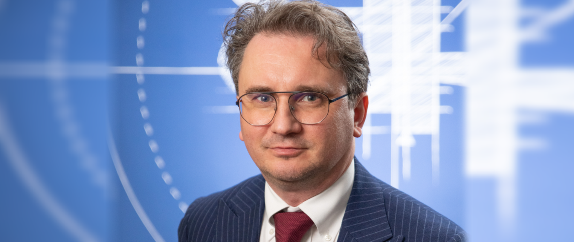 Na niebieskim tle z białym logiem NCBR lekko uśmiechnięty, elegancko ubrany mężczyzna w okularach