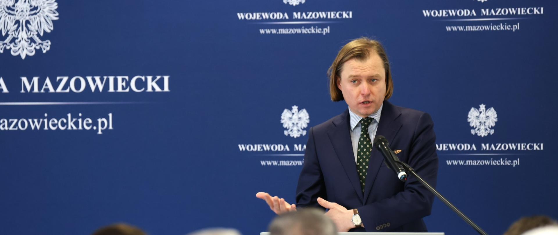 Na zdjęciu mężczyzna przemawia do mikrofonu. Stoi przy szarej mównicy z orłem na przodzie. 