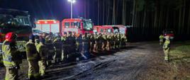 Zbiórka strażaków na podsumowaniu ćwiczeń, w tle samochody pożarnicze. Pora nocna