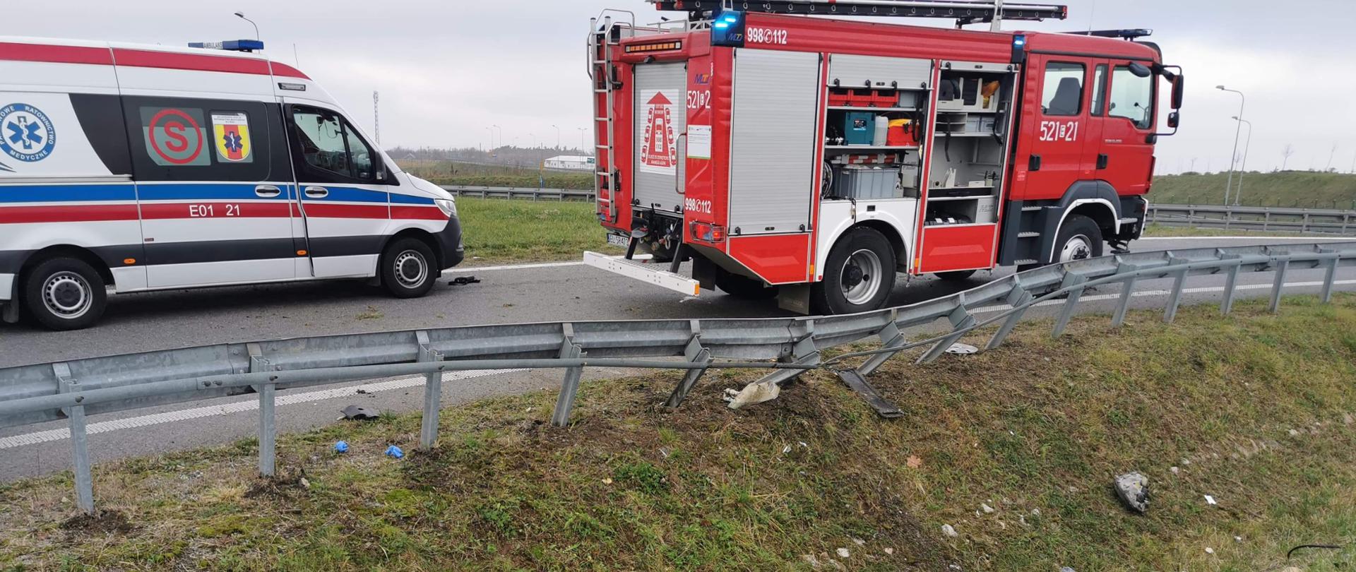 Zdjęcie przedstawia dwa samochody stojące na drodze. Pierwszy samochód to karwtka pogotowia, a drugi to samochód strażacki. Obok samochodów znajduje się rów, w którym widać resztę części samochodu, który miał wypadek. Pomiędzy samochodami a rowem znajduje się barierka ochronna, która została wygięta w kierunku rowu w skutek uderzenia w nią samochodu osobowego.