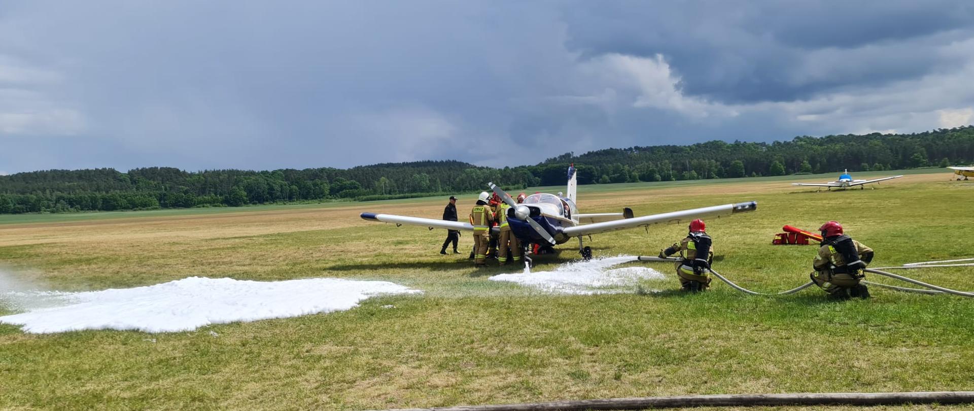 Zdjęcie przedstawia trawiastą płytę lotniska, na której stoi lekki samolot. Wokół niego pracują strażacy w piaskowych mundurach i hełmach. W tle las i wzniesienia.