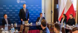 Przy stołach ustawionych w czworokąt siedzi kilka osób, w centralnej części minister Czarnek stoi i coś mówi, za nim niebieska ściana z napisem Ministerstwo Edukacji i Nauki.