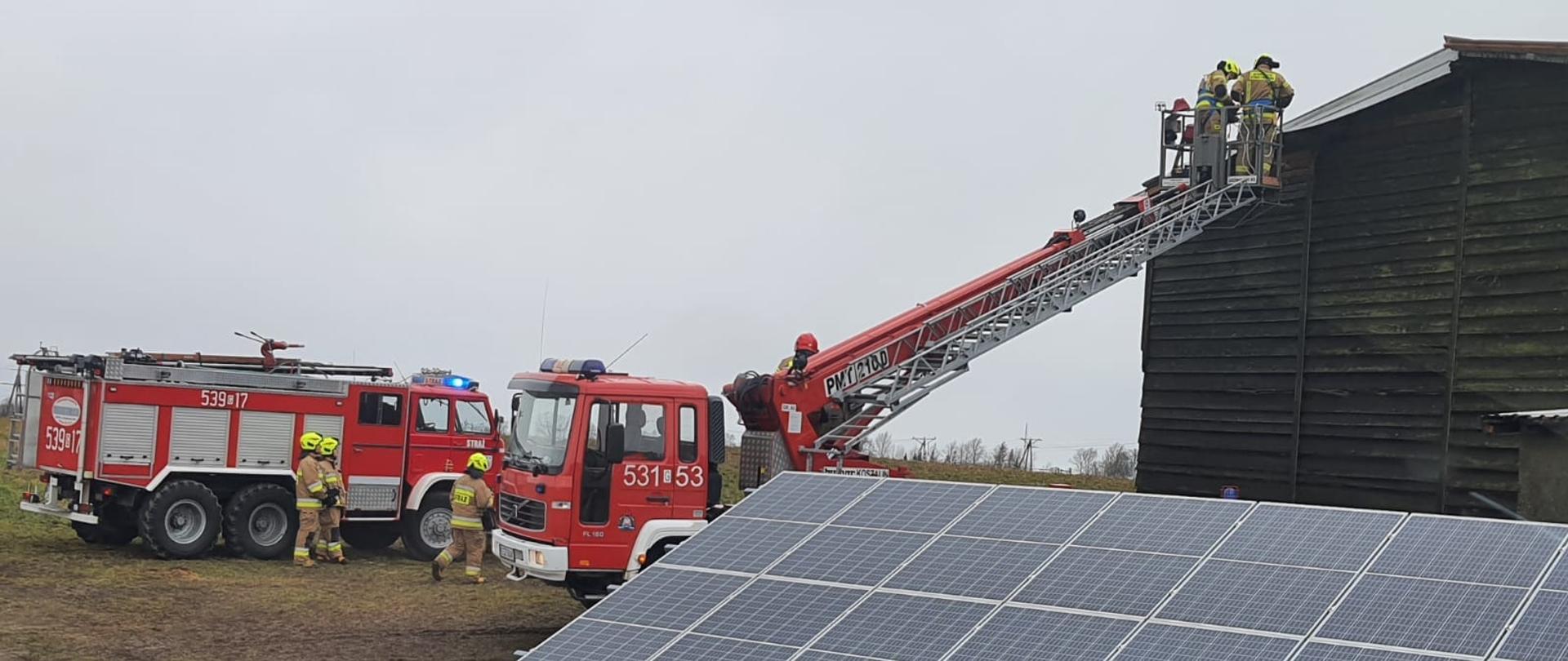 pora dzienna, kilku umundurowanych strażaków stoi miedzy dwoma wozami, gaśniczym i drabina mechaniczną. W koszu drabiny wysuniętym do szczytu stodoły której dach uszkodził silny wiatr znajdują się dwaj ratownicy.