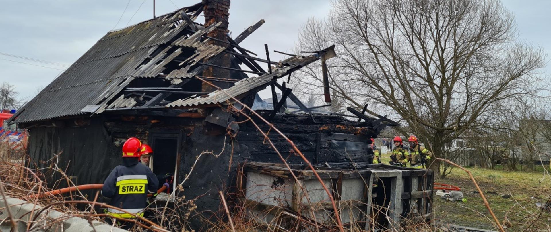 Zdjęcie przedstawia częściowo spalony dom jednorodzinny. Budynek drewniany, kryty eternitem. Część dachu budynku zapadła się na skutek pożaru. Obok budynku znajdują się strażacy ubrani w ubrania specjalne w kolorze ciemnym granatowym oraz żółtym. Na głowach mają czerwone hełmy. W rękach trzymają węże pożarnicze przy pomocy, których dogaszają płomienie. 