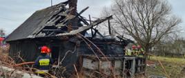Zdjęcie przedstawia częściowo spalony dom jednorodzinny. Budynek drewniany, kryty eternitem. Część dachu budynku zapadła się na skutek pożaru. Obok budynku znajdują się strażacy ubrani w ubrania specjalne w kolorze ciemnym granatowym oraz żółtym. Na głowach mają czerwone hełmy. W rękach trzymają węże pożarnicze przy pomocy, których dogaszają płomienie.