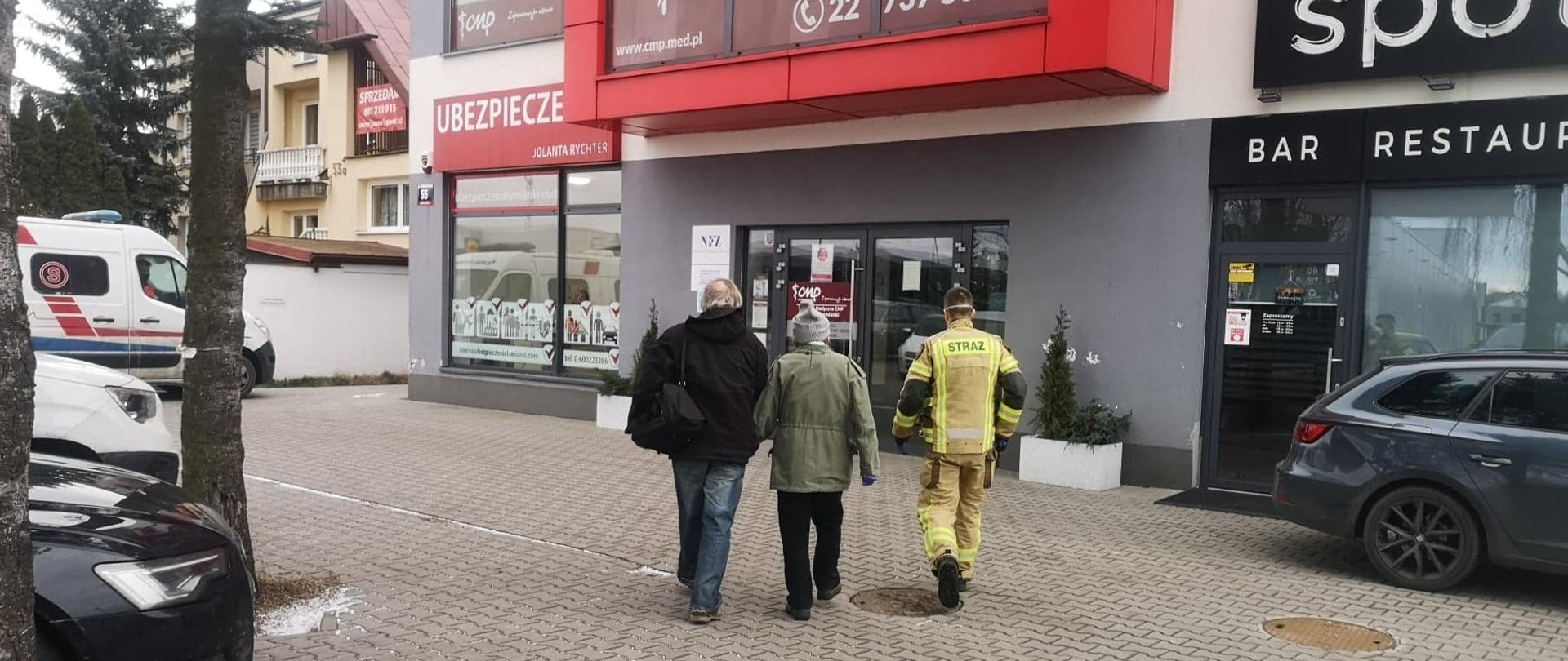 Transport seniorów do punktów szczepień, na zdjęciu senior w asyście opiekuna i strażaka prowadzona do punktu szczepień