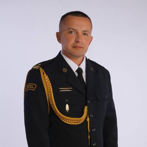 Powyższe zdjęcie to portret mężczyzny w ciemnym, galowym mundurze, prawdopodobnie oficera służb mundurowych.
Mężczyzna ma krótkie, starannie ułożone, jasne włosy i patrzy lekko w bok od obiektywu. Jego twarz jest poważna. Ubrany jest w ciemny mundur z białą koszulą i czarnym krawatem.
Na jego prawym ramieniu widać naszywkę z napisem "PAŃSTWOWA STRAŻ POŻARNA", co sugeruje przynależność do tej formacji. Na lewym ramieniu widoczny jest pagon ze złotą gwiazdką i dwiema belkami (co sugeruje stopień oficerski) oraz ozdobny, złoty pleciony sznur. Na piersi, po lewej stronie, przypięta jest złota plakietka z napisem "P. KNYSAK".