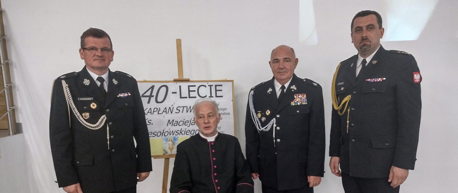 Pomieszczenie, dzień. Na tle białej ściany na krześle siedzi ksiądz ubrany w czarną sutannę z fioletowymi guzikami . Za księdzem stoi sztaluga z tablicą, na której widać częściowo napis "40-lecie kapłaństwa". Z lewej strony księdza stoi druh ubrany w czarny mundur galowy ze srebrnym sznurem galowym bez nakrycia głowy. Z prawej strony księdza stoi druh ubrany w czarny mundur galowy ze srebrnym sznurem galowym bez nakrycia głowy oraz funkcjonariusz PSP ubrany w czarny mundur wyjściowy ze złotym sznurem galowym, bez nakrycia głowy. 