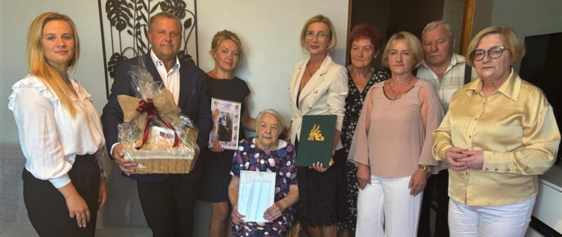 W pomieszczeniu znajduje się grupa stojących osób. Mężczyzna trzyma w ręce kosz prezentowy, stojąca obok kobieta trzyma dyplom. Kolejna kobieta w jasnej marynarce trzyma w ręce ozdobną zieloną teczkę ze złotym logo KRUS. Pomiędzy nimi siedzi Szanowna Jubilatka Pani Stanisława Cebulak, która trzyma w rękach upominek. Obok stoją członkowie rodziny jubilatki. Przed nimi stoi duży kosz jasnych kwiatów.