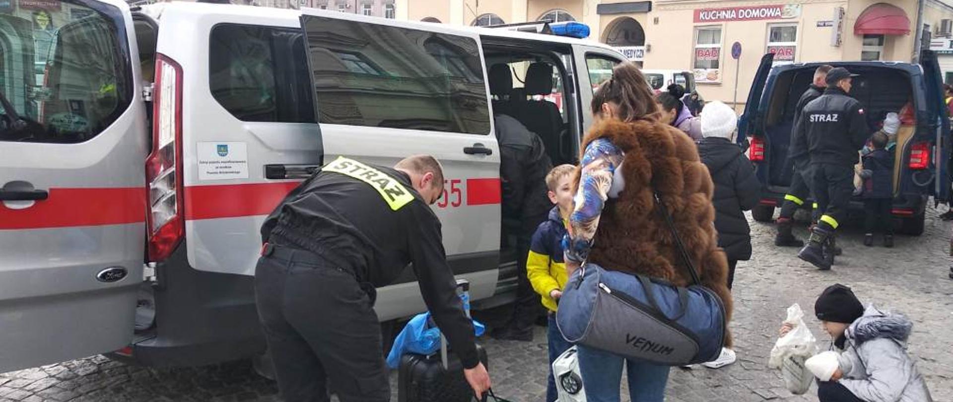 Strażacy KP PSP w Brzozowie pomagają uchodźcom z Ukrainy. Fotografia wykonana w jasny dzień, na której widać dwóch umundurowanych strażaków na tle srebrnego pojazdu pożarniczego. Jeden pomaga przenosić bagaż stojącym obok osobom cywilnym, drugi wyciąga bagaże z busa. W głębi inne pojazdy, strażacy i osoby cywilne. W tle widoczne ściany budynków miejskich.