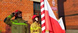Na zdjęciu widać poczet flagowy składający się z trzech strażaków, którzy zaczynają wciągać Flagę Państwową na maszt. W tle widać budynki strażnicy z czerwonej cegły