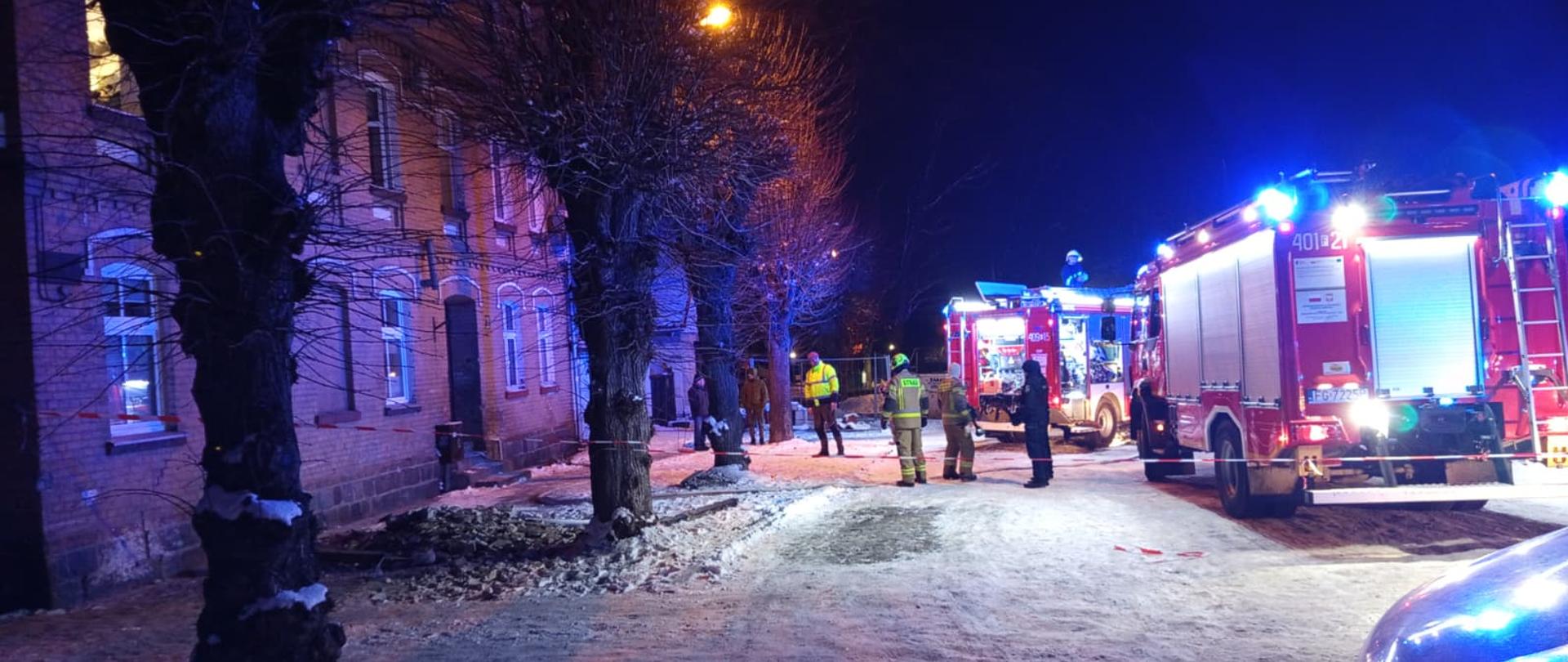 Fotografia przedstawia ujęcie przed kamienicą, jest pora nocna, teren zaśnieżony, przed kamienicą stoją pojazdy gaśnicze straży pożarnych, widoczni są strażacy, teren jest oświetlany.