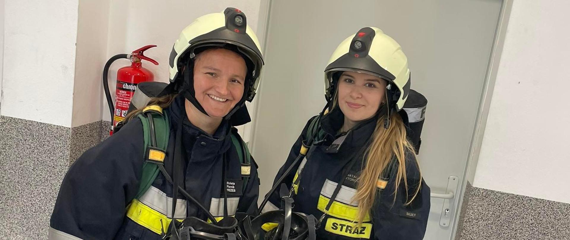 Dwie druhny przed wejściem do komory dymowej w ubraniach specjalnych, aparatach powietrznych i hełmach.