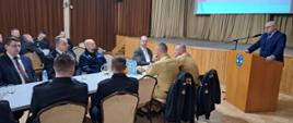 Strażacy ubrani w granatowe mundury galowe, oraz pisakowe mundury siedzą przy stolikach, w tyle rolap z logiem Państwowej Straży Pożarnej na tylnej ścianie kotary oraz ekran z wyświetloną prezentacją. Przy mównicy stoi starosta ubrany w garnitur, przed sobą ma mikrofon