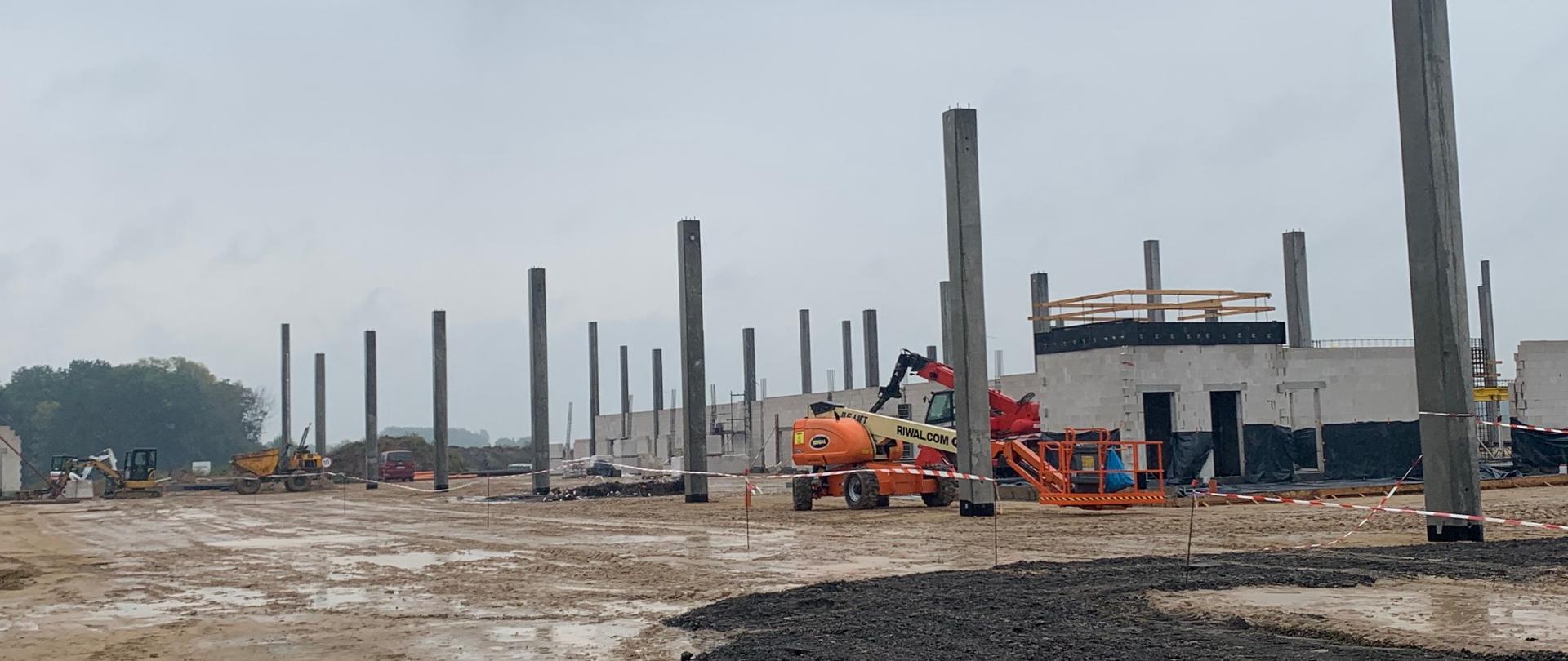 Zdjęcie przedstawia teren rozbudowy terminala Portu Lotniczego Radom-Sadków. Na pierwszym planie ciemny żwir i piach oraz wbite w ziemie betonowe pale. W tle niska zabudowa obiektów tworzących integralną część terminala