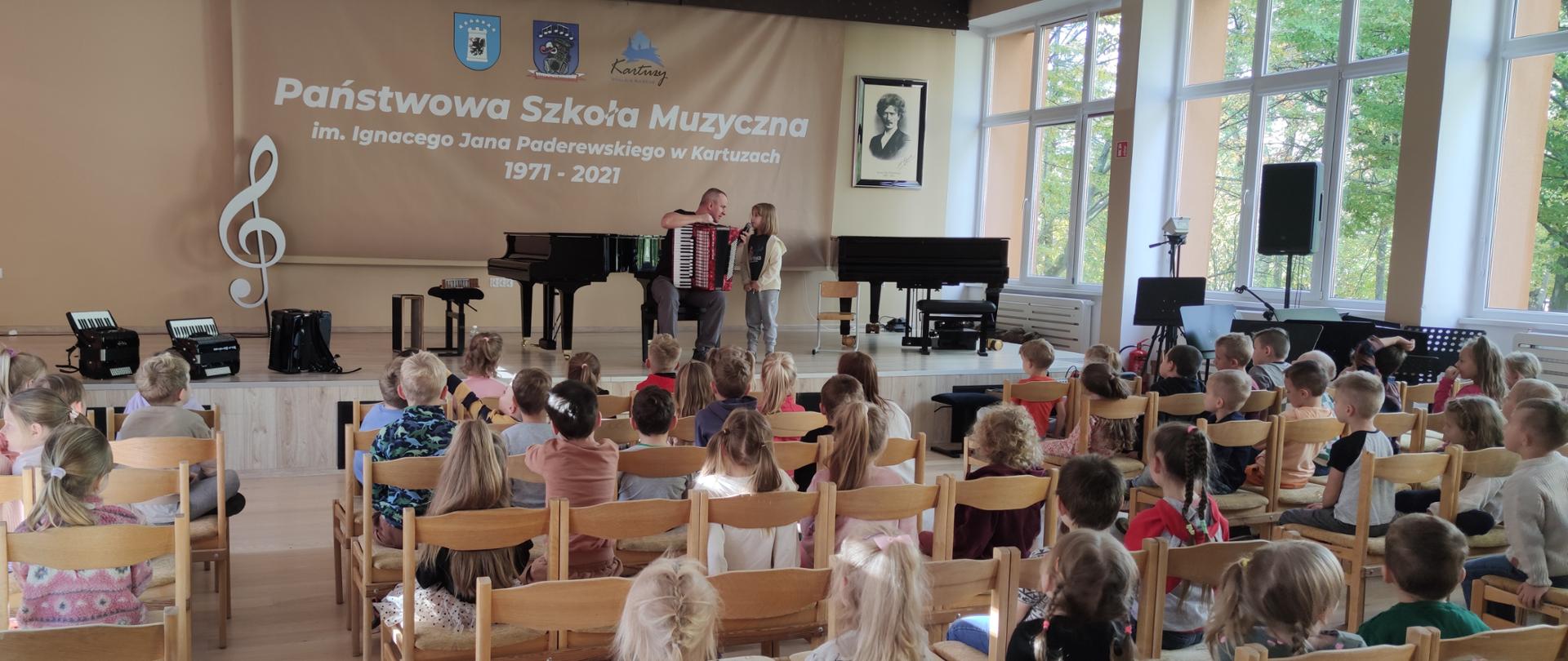 na scenie nauczyciel z akordeonem, na widowni dzieci z przedszkoli