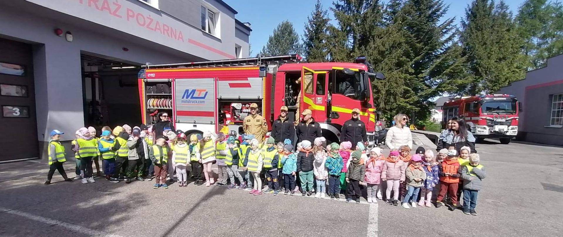 Dzieci z wizytą w Państwowej Straży Pożarnej. W tle samochód strażaki. Dzieci kolorowo ubrane. Słonecznie.