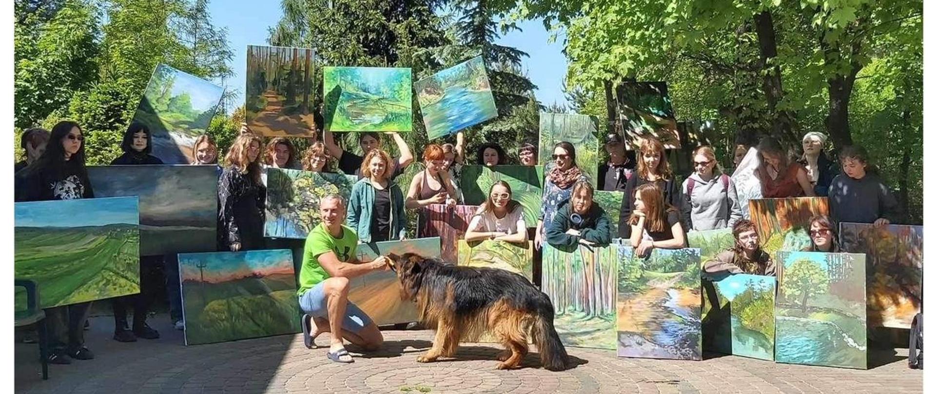 Zdjęcie grupowe. Słoneczna pogoda. Część uczniów trzyma w rękach obrazy w powietrzu lub podparte o podłoże. W grupie znajduje się nauczyciel Anna Chmielnik. Na przodzie nauczyciel Tomasz Krasowski ukląkł na jednym kolanie i trzyma przedmiot w pysku owczarka niemieckiego. W tle bujna roślinność, drzewa.