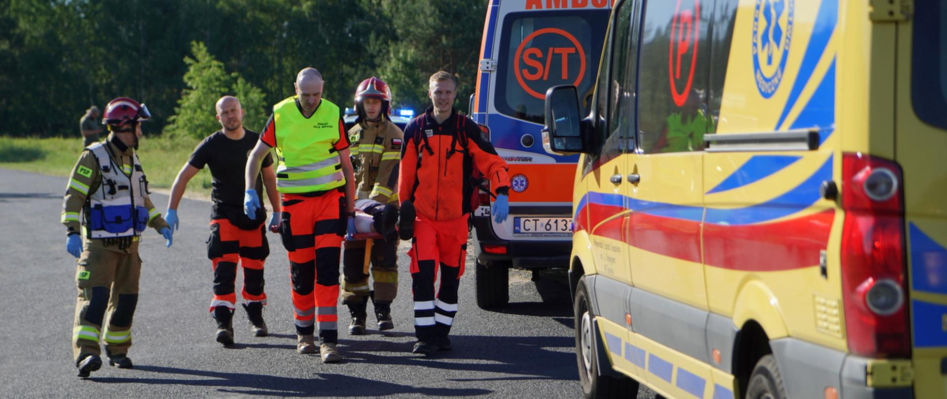 Zespół ratownictwa medycznego oraz strażacy Państwowej Straży Pożarnej wspólnie transportują osobę poszkodowaną. Idą asfaltową drogą, niosąc pacjenta na miękkich noszach. Po prawej stronie kadru stoją dwa żółte ambulanse, z których jeden ma widoczną rejestrację z Torunia (CT 6133). W tle widać zieloną ścianę lasu w słoneczny dzień.