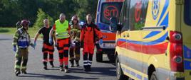 espół ratownictwa medycznego oraz strażacy Państwowej Straży Pożarnej wspólnie transportują osobę poszkodowaną. Idą asfaltową drogą, niosąc pacjenta na miękkich noszach. Po prawej stronie kadru stoją dwa żółte ambulanse, z których jeden ma widoczną rejestrację z Torunia (CT 6133). W tle widać zieloną ścianę lasu w słoneczny dzień.