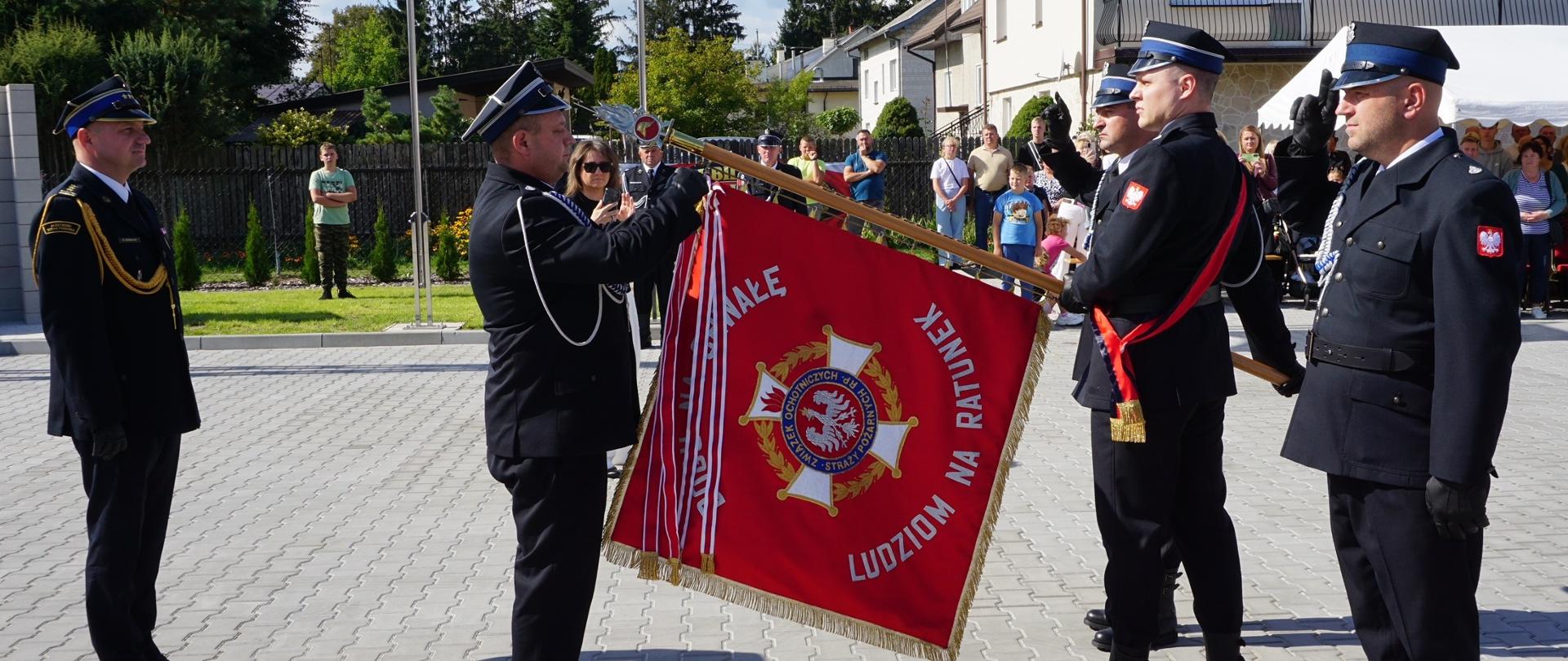 Poczet sztandarowy Ochotniczej Straży Pożarnej w Krypach uczestniczy w ceremoniale podczas obchodów 100-lecia jednostki w czasie odznaczania. Strażacy stoją przy sztandarze organizacji, a uroczystości przyglądają się mieszkańcy i zaproszeni goście.