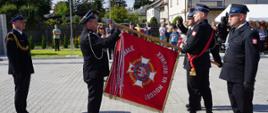 Poczet sztandarowy Ochotniczej Straży Pożarnej w Krypach uczestniczy w ceremoniale podczas obchodów 100-lecia jednostki w czasie odznaczania. Strażacy stoją przy sztandarze organizacji, a uroczystości przyglądają się mieszkańcy i zaproszeni goście.