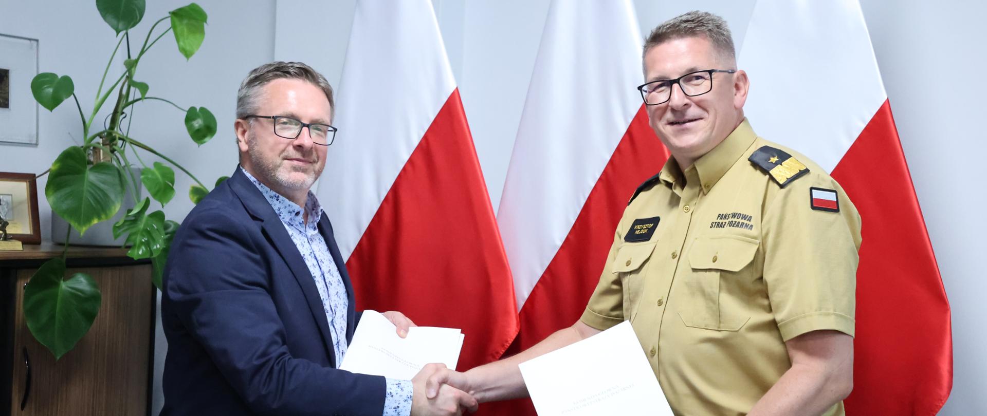 Zdjęcie pamiątkowe uczestników spotkania. Dwaj mężczyźni prezentujący podpisane dokumenty w teczkach, ściskający sobie dłonie. W tle biało-czerwone flagi