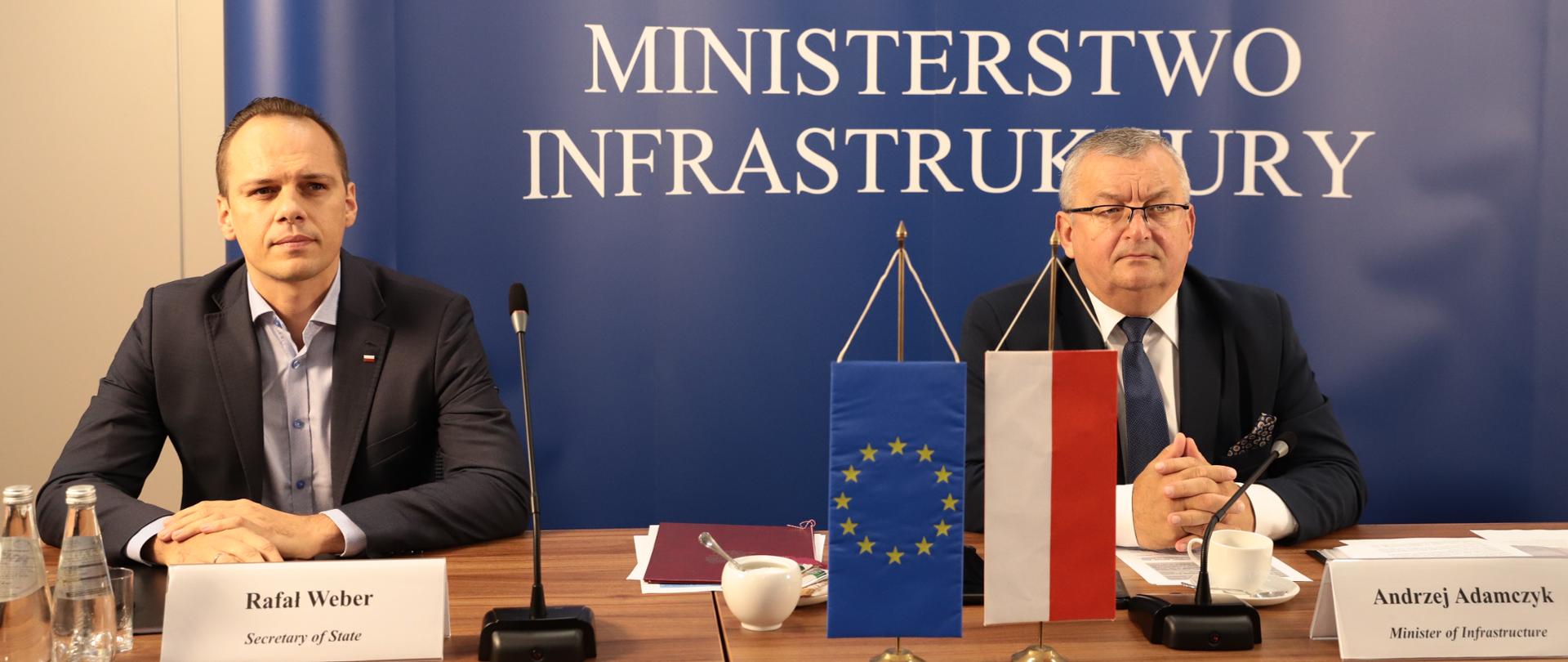 Na zdjęciu widać dwóch mężczyzn siedzących przy stole. W ich tle jest tło z napisem Ministerstwo Infrastruktury. Mężczyźni są ubrani w ciemne garnitury, białe koszule. 