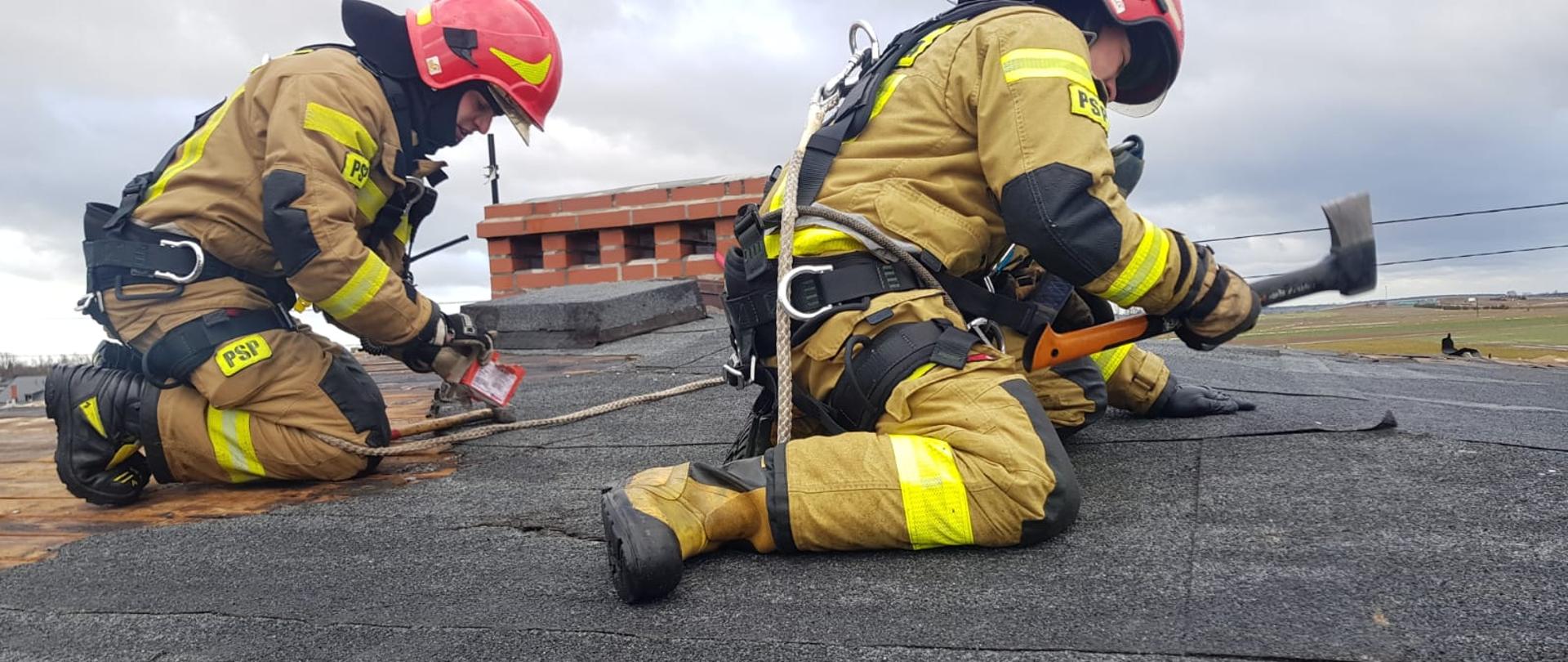 Dwóch strażaków w ubraniu specjalnym w hełmach klęczy i przybija papę na dachu.