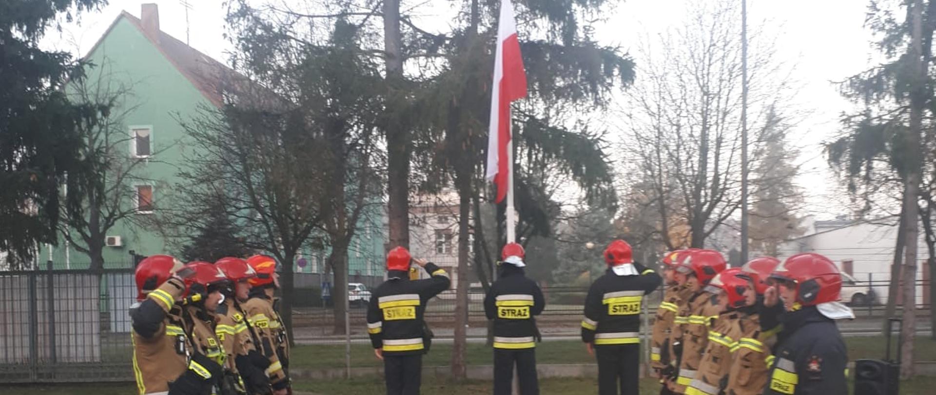Stojący umundurowani strażacy w dwóch szeregach twarzami do siebie. W tle maszt z zawieszoną na nim flagą państwową.