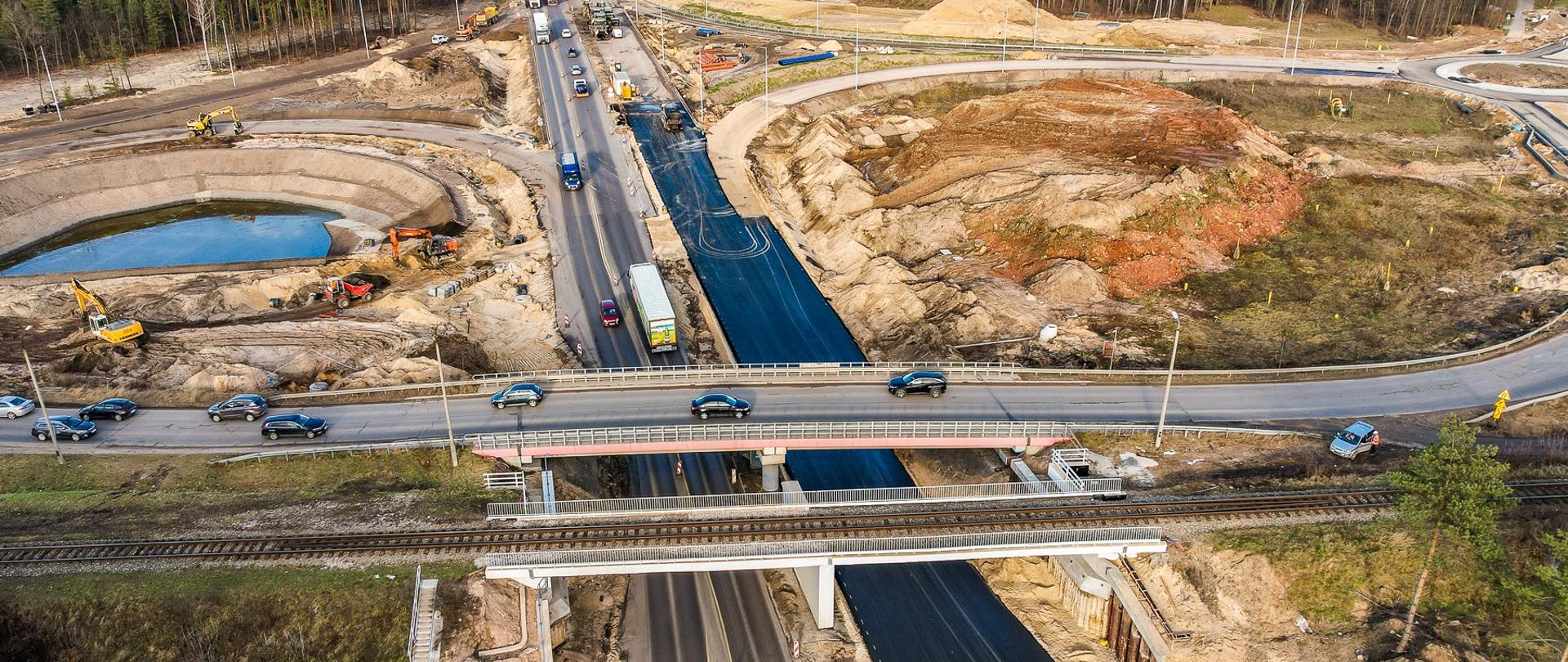 Fotografia przedstawiająca budowany węzeł Dąbrowa Górnicza Pogoria. Na pierwszym planie widoczny dwa obiekty inżynierskie pierwszy kolejowy drugi drogowy. W dalszym planie widoczne maszyny budowlane w trakcie budowania łącznic z węzła.