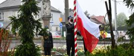Trzech mężczyzn ubranych w czarny mundur wyjściowy strażacki, czarnymi butami, na głowie maja rogatywki, na twarzy ma ubrana maseczkę chirurgiczną koloru biało niebieskiego, stoją na zewnątrz w pobliżu masztu. Dwóch mężczyzn podtrzymuje flagę państwową i zapina ja do sznurka na maszcie. Jeden mężczyzna stoi w odległości 2 metrów i salutuje. W tle jest figurka przydrożna oraz drzewka i krzewy. Z tyłu w tle znajduje się budynek koloru szarego
