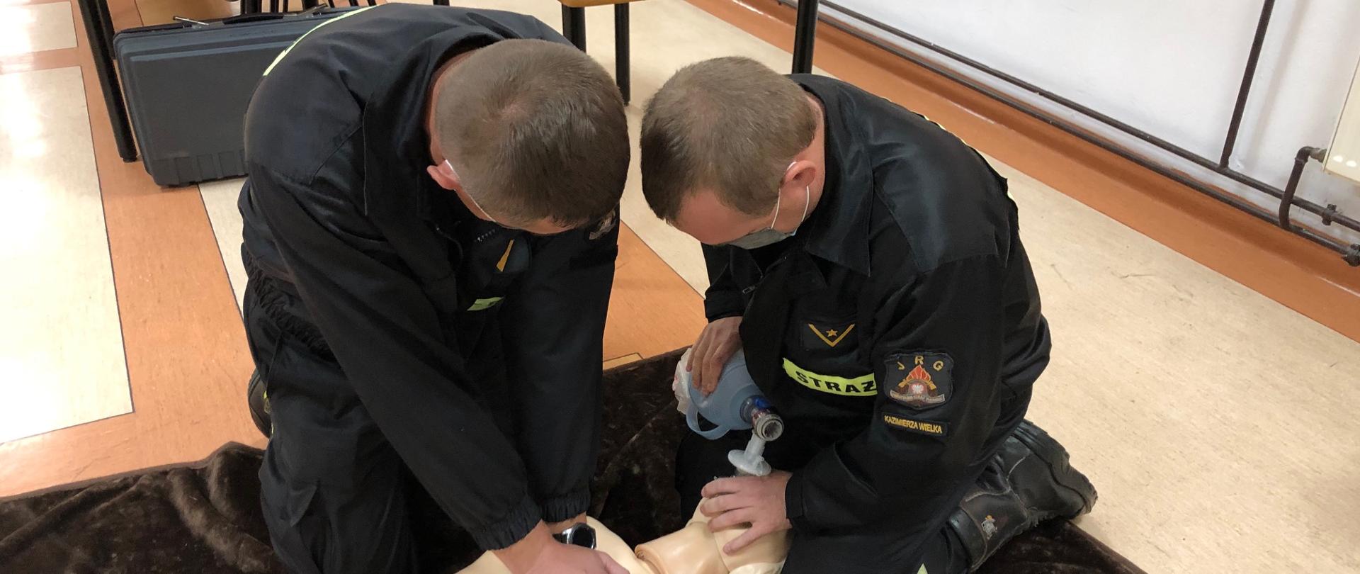 Na pierwszym zdjęciu strażacy-ratownicy wykonują symulację resuscytacji krążeniowo-oddechowej u osoby dorosłej. Jeden z ratowników wykonuje 30 uciśnięć klatki piersiowej, natomiast drugi wykonuje 2 oddechy ratownicze za pomocą worka samorozprężalnego.