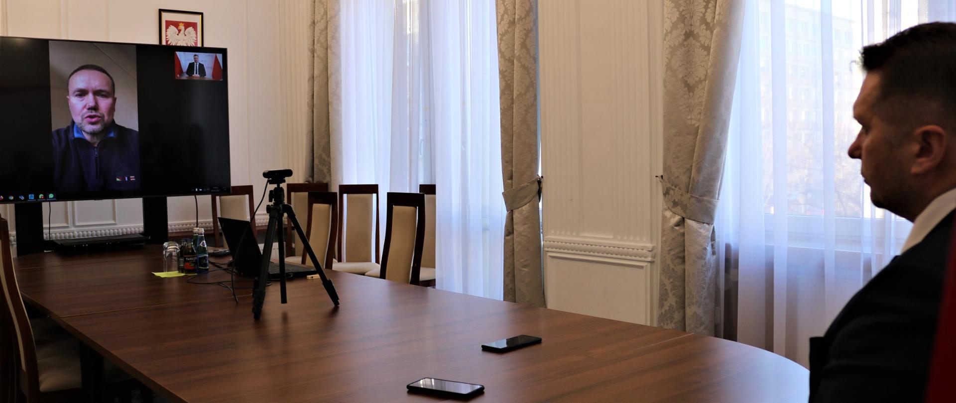 Duży ekran telewizyjny, ktory stoi na końcu stołu. Na monitorze w prostokącie widoczny mężczyzn ubrany w grantowa bluzę. Po drugiej stronie stołu minister Przemysław Czarnek. Widoczny jest tylko fgrmant postaci. na stole lężą telefony i, stoi laptop i sprzęt do nagrywania. W tle jasne załsony i firany i krzesła stojące w rogu. 