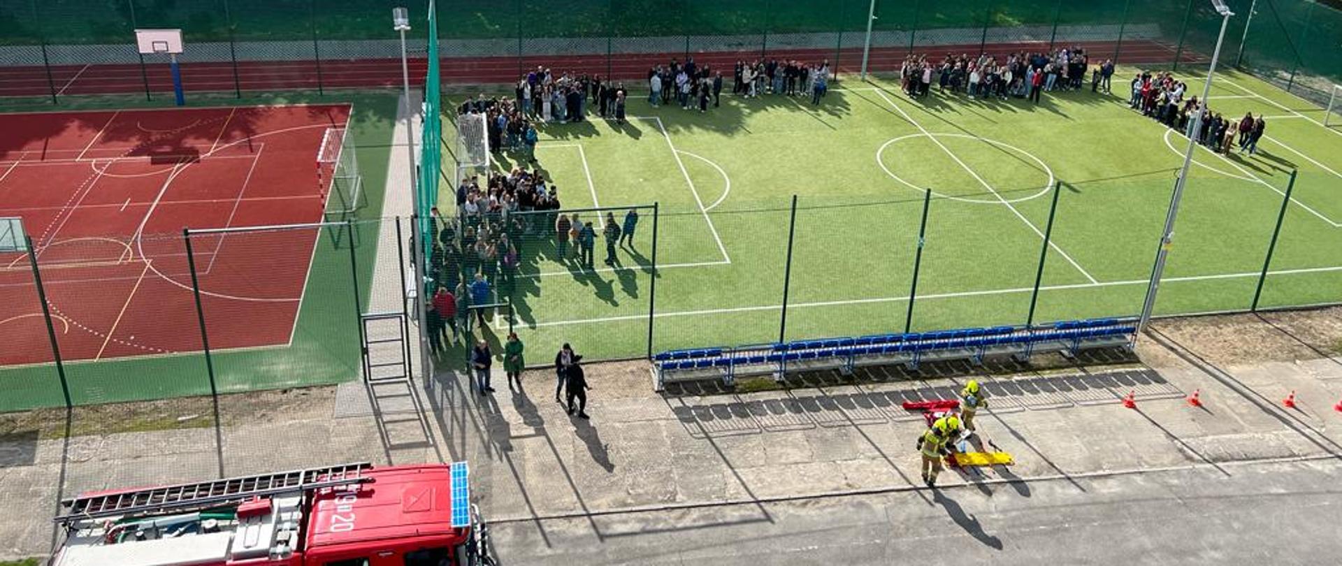 Zdjęcie zrobione z kosza podnośnika hydraulicznego. Na boisku sportowym znajdują się ewakuowani wcześniej uczniowie oraz pracownicy szkoły. Widać też kilku strażaków oraz pojazd pożarniczy.