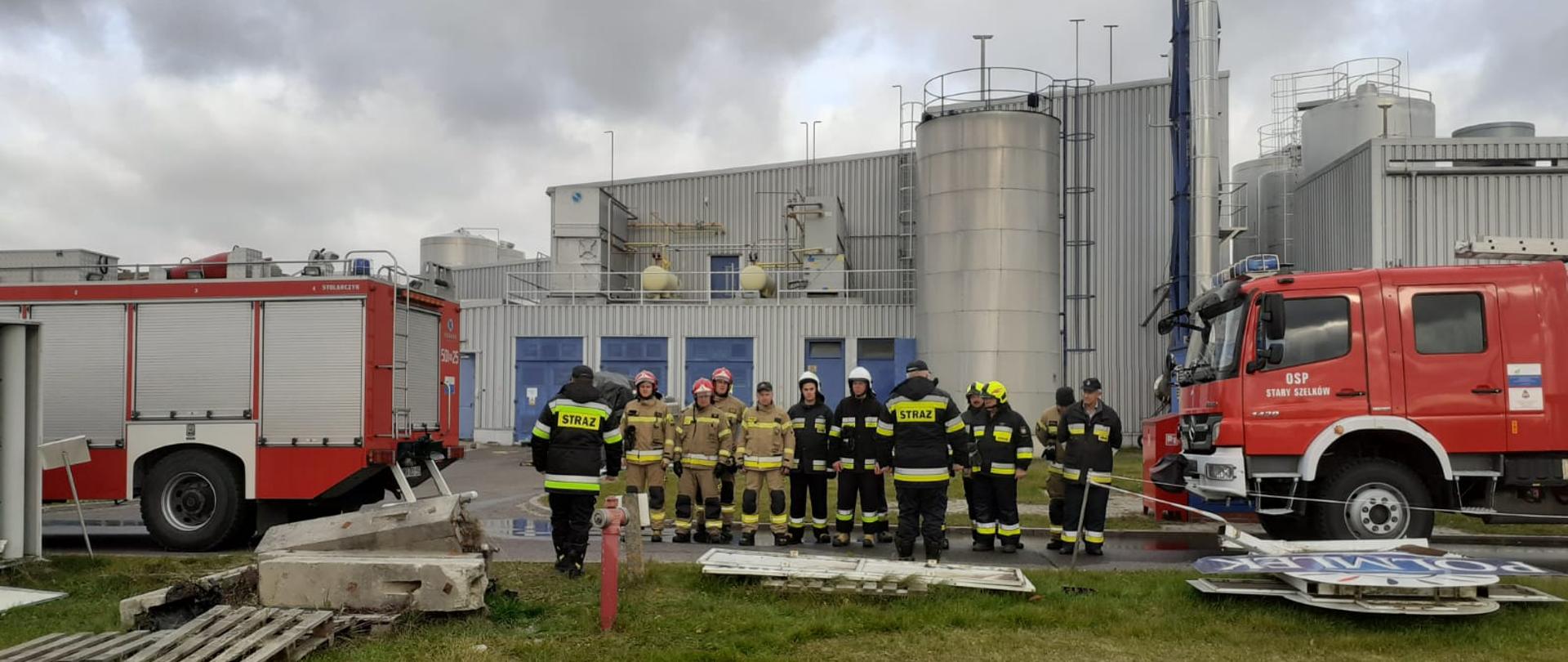 Strażacy stoją na zbiórce przed zakładem, pomiędzy samochodami strażackimi. Kierownik ćwiczeń oraz rozjemca stoją przed nimi i omawiają ćwiczenia.