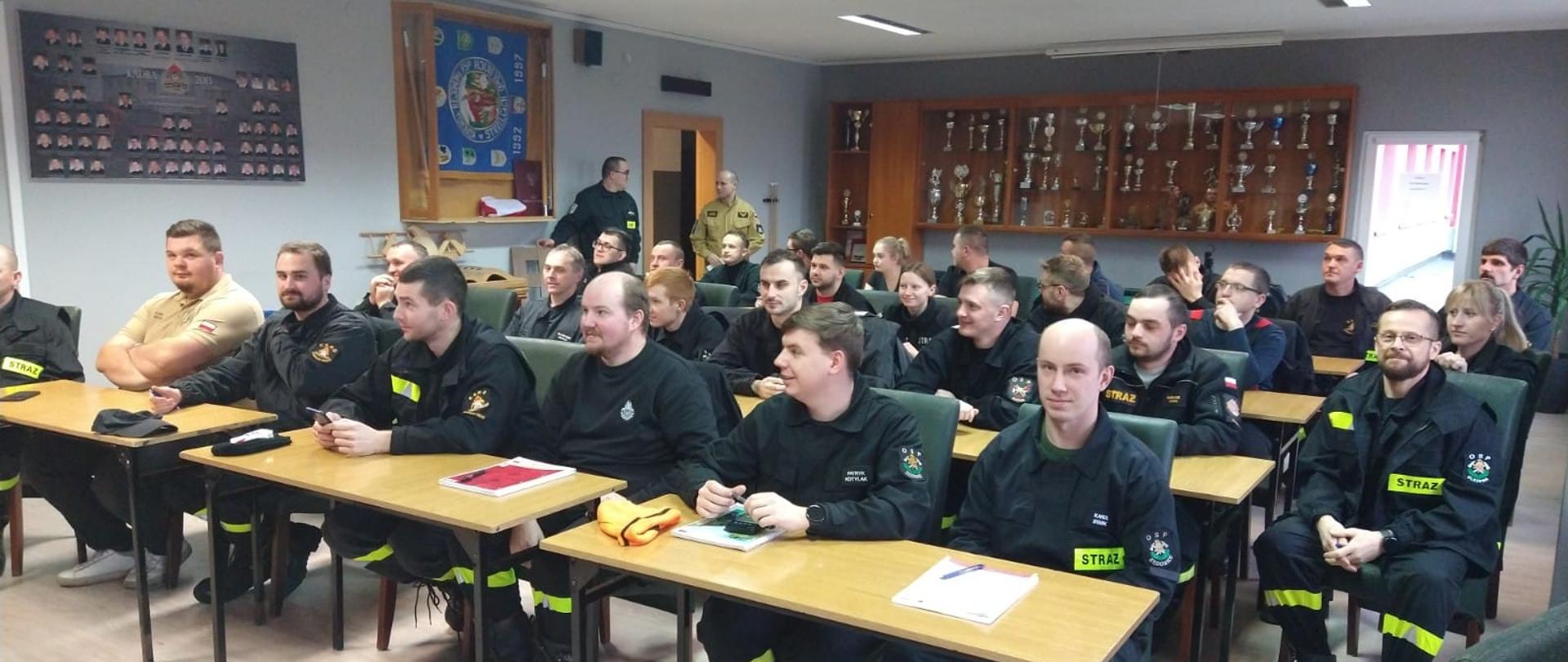 Na zdjęciu znajdują się druhowie OSP siedzący za ławkami na sali wykładowej, w tle stoi strażak
