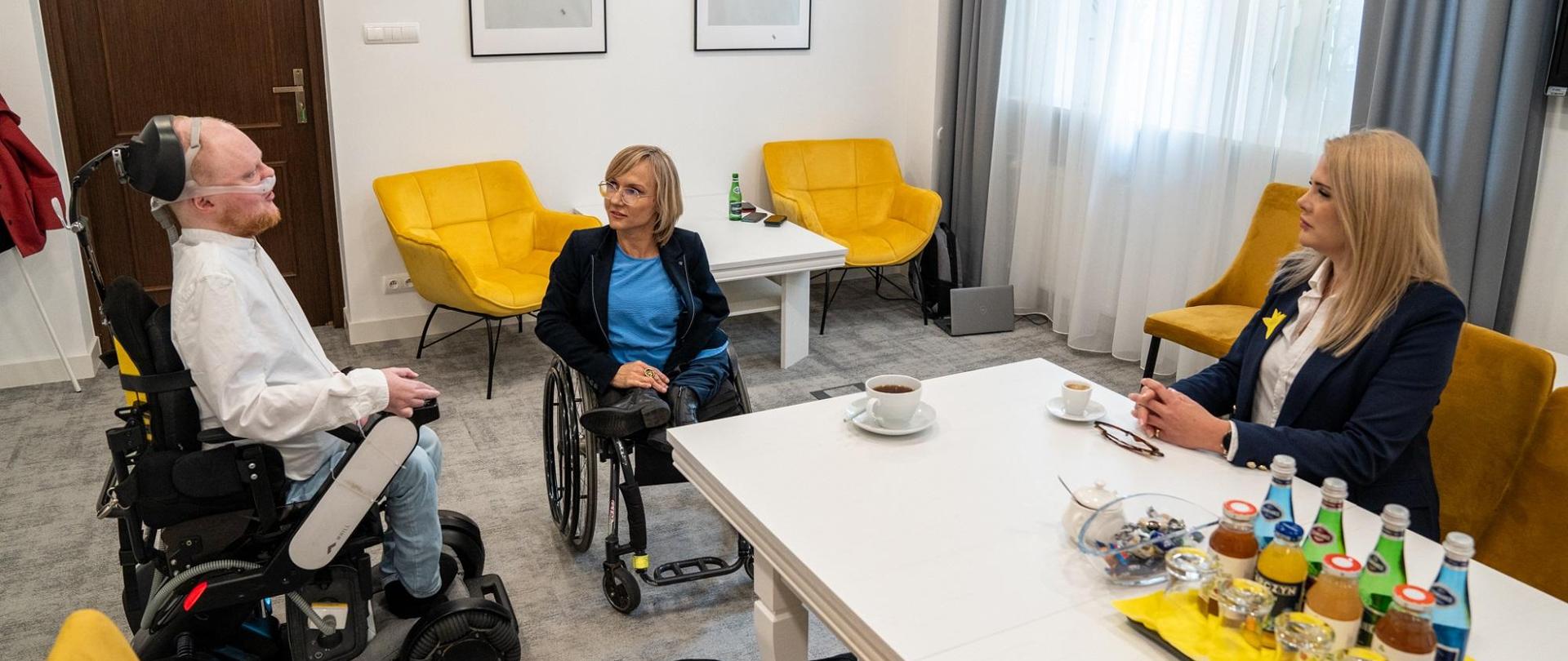 W pokoju z białymi ścianami za stołem siedzi dyrektor Matusik, rozmawia z mężczyzną w białej koszuli i kobietą w czarnej marynarce, oboje siedzą na wózkach inwalidzkich.