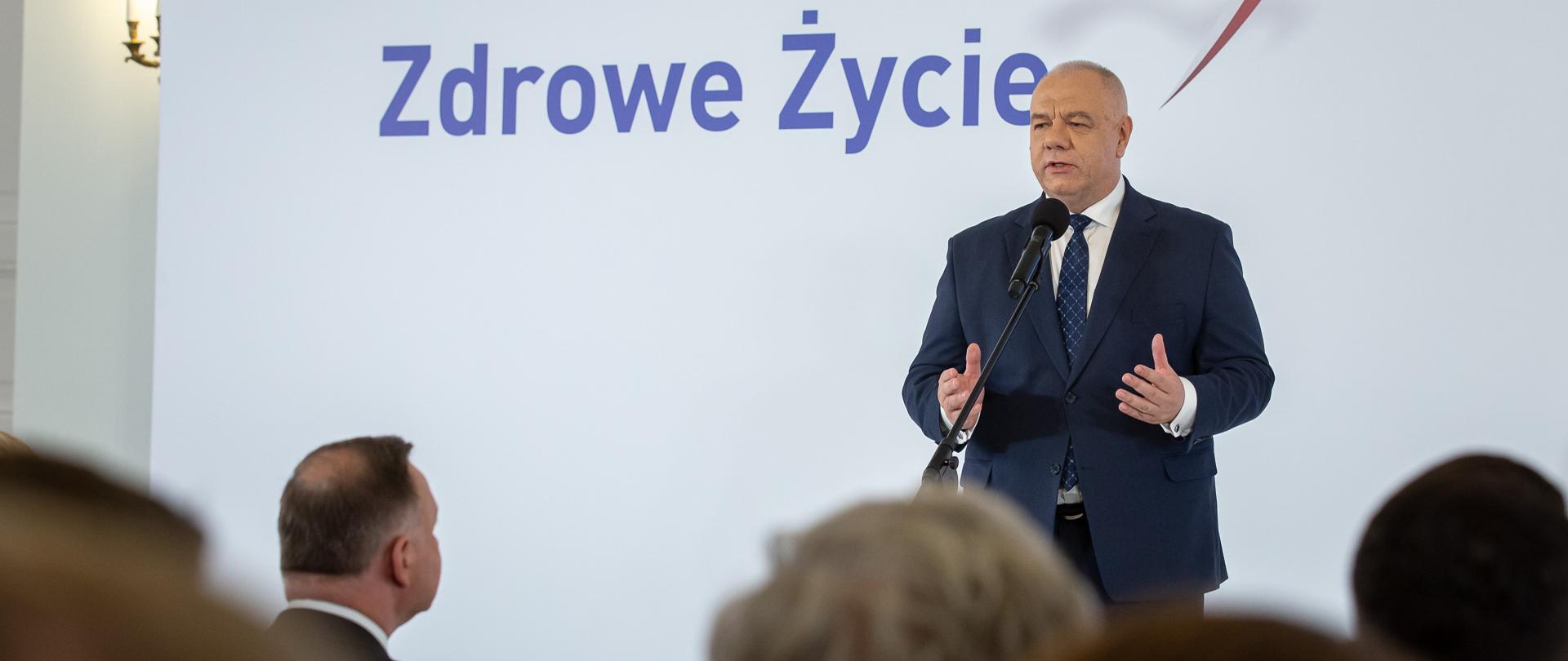 Wicepremier Jacek Sasin stoi za mikrofonem. W tle ścianka z napisem Zdrowe Życie. 