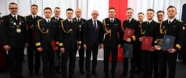 Pamiątkowe zdjęcie grupowe przedstawicieli kierownictwa PSP oraz wyróżniających się absolwentów Akademii Pożarniczej, trzymających dyplomy ukończenia studiów. W tle biało-czerwona flaga.